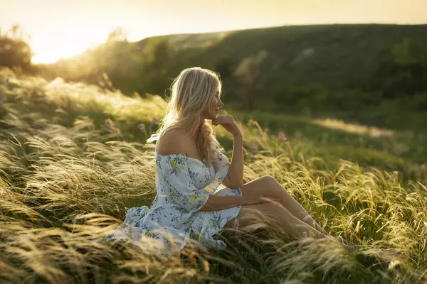 A gentle woman in a dress sits, thinking among the feather grass on the hillside in the gentle sunset light