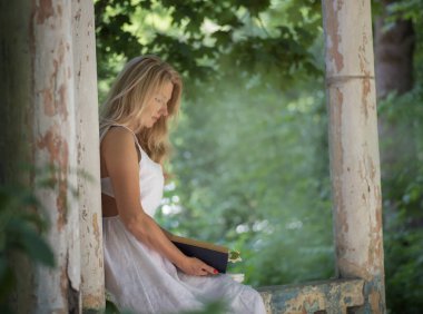 Beyaz elbiseli, uzun sarı saçlı bir kadın bahçedeki çiçekli yasemin çalıları arasında barok sütunları olan eski bir çardakta kitap okuyor..