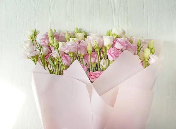 A bouquet of delicate pink and white eustoma lisianthus flowers in pink paper on a white wooden background