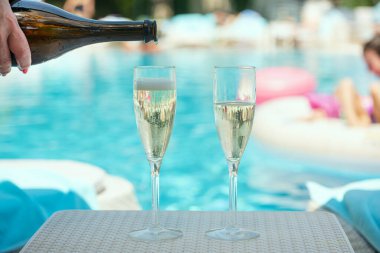 İki bardak şampanya ve parlak mavi bir yüzme havuzunun yanındaki şişeden su dolduran bir el. Su kenarında gevşeme.