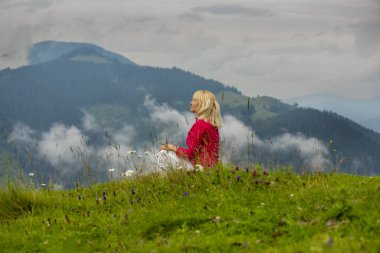 Orta yaşlı sarışın bir kadın dağların tepesindeki bir açıklıkta oturuyor. Yabani çiçekler ve bulutların arasında. Bir anlık zevk..