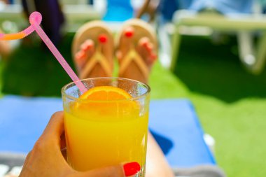 Bir bardak portakallı limonata ve elinde bir dilim buz ile arka planda kırmızı manikürlü havuz kenarındaki plaj terlikleri.