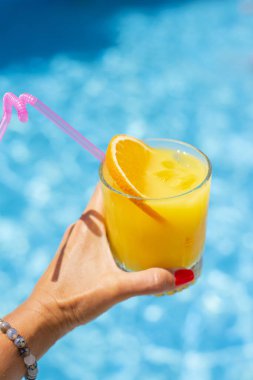 Elinde bir dilim portakal ve buz olan bir bardak portakallı limonata mavi havuz suyunun arka planında. 