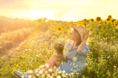  Elinde papatya buketi ve hasır şapkalı bir kadın gün batımında çiçeklerle ve ayçiçekleriyle dolu bir tarlanın tadını çıkarıyor. Gerçekten basit bir kadın. Model değil, rötuş yapmadan.