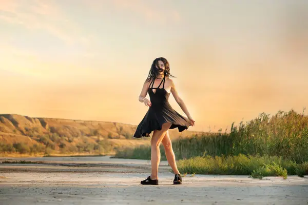 Stock image Girl with black long curly hair and a short black dress has fun happily spinning on the shore of a reservoir among wild shores. 