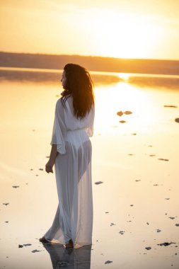 Uzun siyah kıvırcık saçlı, uzun beyaz elbiseli güzel bir kız gün batımında gölün suyunda duruyor. Göl yüzeyindeki gökyüzünün yansıması. İnanılmaz güzel manzara. 