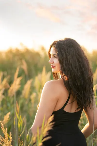 stock image  A girl with dark curly hair in a short dress among the grass on the shore of a pond at sunset. 