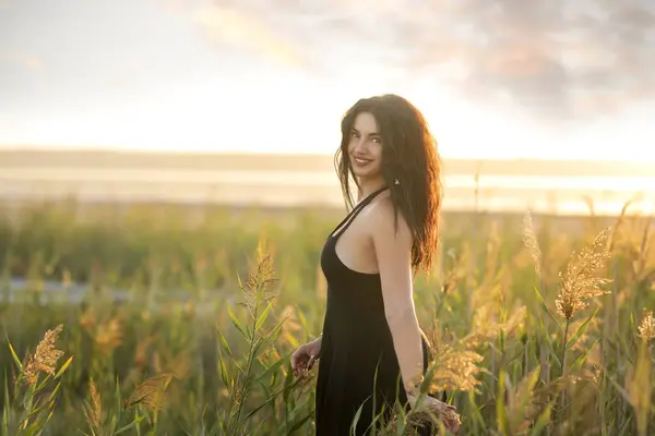 stock image  A girl with dark curly hair in a short dress among the grass on the shore of a pond at sunset. 