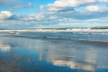 Dalgalarla kaplı deniz manzarası ve kıyı şeridinin suyundaki bulutların yansıması..