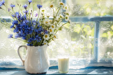 Bir buket çiçek ve seramik sürahide papatyalar ve tahta bir pencerede bir bardak süt. rustik biçim