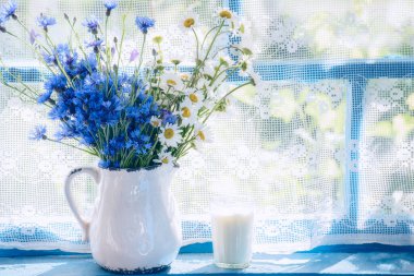  Seramik bir sürahide bir buket çiçek ve papatya ve tahta bir pencerede bir bardak süt. ülke tarzı 