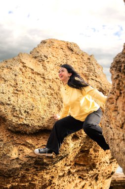 Sarı kazaklı ve siyah pantolonlu genç bir kadın kayalıklardan atlıyor, gülümsüyor. Rüzgâr siyah saçlarını savururken dramatik bir gökyüzüne karşı maceraperest bir açık hava anının tadını çıkarıyor.