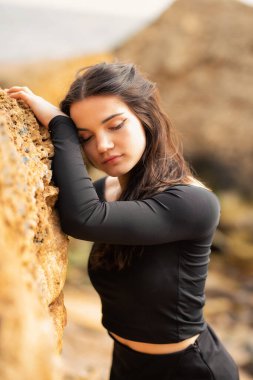 Uzun siyah saçlı genç bir kız arka planda deniz olan bir kayanın yanında poz veriyor. Uzun kollu siyah dar bir bluz giyiyor. Zarif ve sofistike bir görüntü yaratıyor. Onun delici bakışları...