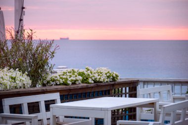 Güneşin doğuşunda huzurlu bir okyanus manzaralı büyüleyici bir sahil kafesi. Beyaz ahşap masalar ve çiçek açan çiçekler deniz kenarında mükemmel bir sabah için samimi ve romantik bir ortam yaratır..