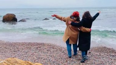 İki kız arkadaş soğuk deniz kıyısında yürüyor, sarılıyor ve uzaklığa bakıyor. Serin bir günde dostluk ve beraberlik anı..