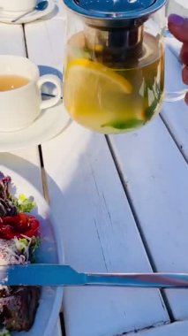 Peynirli salatalı bir tabak, şeffaf aromalı çaydanlık aromalı bir çaydanlıktan beyaz ahşap bir masada portakal ve nane ile yavaşça dökülen bir kadın eli. yumuşak sabah güneşi