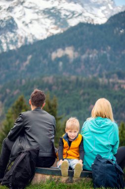 Bir bankta oturan aile, kameraya arkası dönük, dağ manzarasına bakıyor. Sarı yelekli çocuk kameraya gülümsüyor. Bahar günü, kar zirveleri, orman, huzurlu manzara..