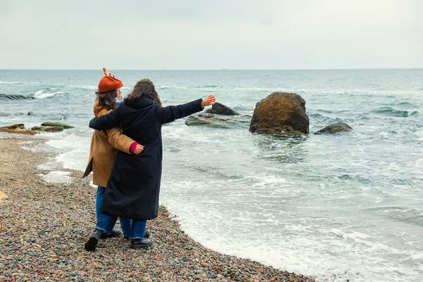 İki kadın, yağmurlu ve soğuk bir deniz kıyısında, soğuk ve bulutlu havaya rağmen, sıcak bir kucaklaşma ve neşeli bir anı paylaşarak kol kola dururlar..