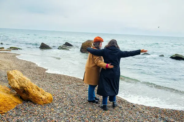 İki kadın, yağmurlu ve soğuk bir deniz kıyısında, soğuk ve bulutlu havaya rağmen, sıcak bir kucaklaşma ve neşeli bir anı paylaşarak kol kola dururlar..