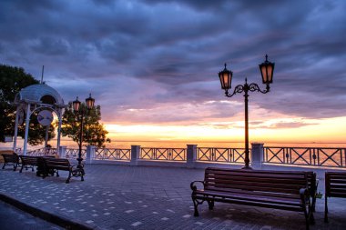 Gün doğumunda banklar ve fenerlerle kıyı yürüyüşü. Romantik gökyüzü ile sakin deniz manzarası, dramatik bulutlar, ve yumuşak ışık mükemmel sakin sabah sahnesi.