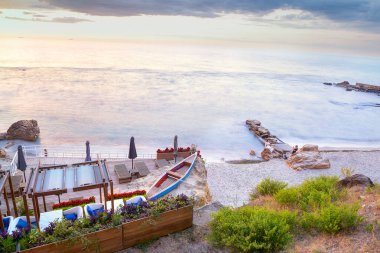 Şafak vakti yeşil taşlı sakin bir sahil. Bulanık su, uzun süre maruz kalma, yumuşak güneş ışığı. Mükemmel bir sabahın huzurlu manzarası. Kızlar kayaların üstünde fotoğraf çekiyor..