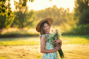 Yaz elbisesi ve hasır şapkalı genç bir kadın gün batımında güneşli bir çayırda bir buket kır çiçeği tutuyor. Romantik kırsal yaşam tarzı, doğa güzelliği ve mutluluğu..