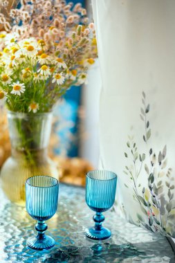 Cam bir masada iki mavi şarap kadehi, taze papatya çiçekli vazo ve perdenin içinden gün ışığıyla rahat bir köy hayatı. Yaz kırsal atmosferi, romantik ev dekorasyonu ve kulübe estetiği.