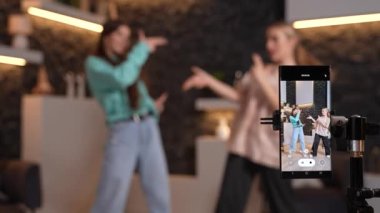 Kadın gençler evde dans ediyor, akıllı telefon kamerasıyla sosyal medya için bir dans videosu kaydediyorlar. Genç blogcular arka planda bulanıklaşıyor ve kanalları için içerik oluşturuyorlar. Yüksek kaliteli görüntüler.