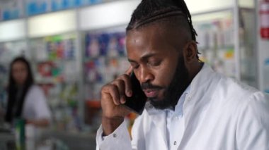 Bu 4K yüksek kaliteli stok görüntülerinde koyu tenli bir eczacı müşteriye cep telefonuyla danışıyor ve bir eczanede dizüstü bilgisayar kullanıyor. Sağlık hizmetlerine müşteri merkezli yaklaşım 