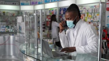 Bir perakende eczanesinde koruyucu sağlık maskesi takan koyu tenli bir erkek eczacının cep telefonu ve dizüstü bilgisayarla müşteriye sağlık danışmanlığı yaptığı 4K yüksek kaliteli görüntüleri.