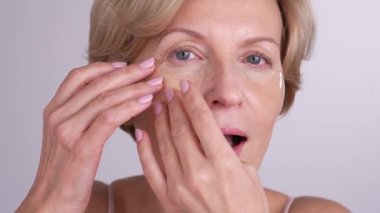 Orta yaşlı bir kadın aynaya bakıyor, göz bandı yapıştırıyor ve gülümsüyor. Zarif yaşlanan dişi, cildine ve güzelliğine özen gösterir.