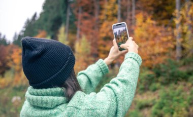 Sıcak siyah şapkalı genç bir kadın, sonbahar ormanının fotoğrafını akıllı bir telefonla çekiyor. Seyahat kavramı..