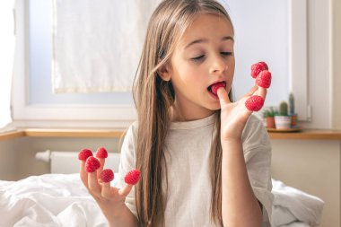 Parmaklarında ahududu olan komik küçük kız sabahları yatakta oturuyor, sağlık ve vitamin kavramı..