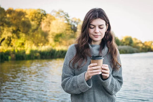 A young woman in a sweater with a cup of coffee on a blurred background of the river.