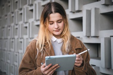 Kadife ceketli çekici genç bir kadın dijital tablet üzerinde kalemle şehirdeki geometrik beton bir duvarın bulanık arka planına çiziyor...