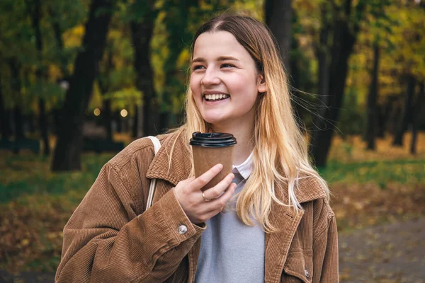 An attractive young woman in a corduroy jacket with a cup of coffee walks in the autumn park.
