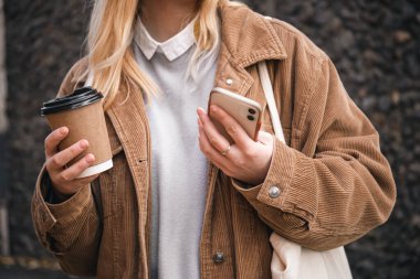 Yakın plan, bir fincan kahve ve kadife ceketli şık bir kadının elinde bir akıllı telefon..