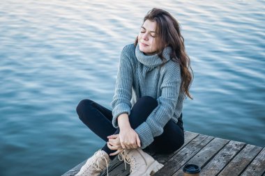 Suyun arka planında, soğuk mevsimde, süveter giyen çekici genç bir kadın nehrin kenarında oturur..