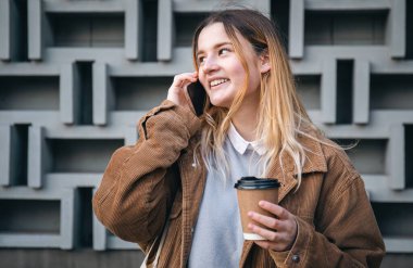Elinde bir fincan kahveyle telefonda konuşan çekici genç bir kadın geometrik beton arka planda....