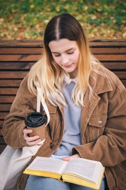 Çekici genç bir kadın sonbahar parkında ahşap bir bankta oturuyor, kahve içiyor ve kitap okuyor..