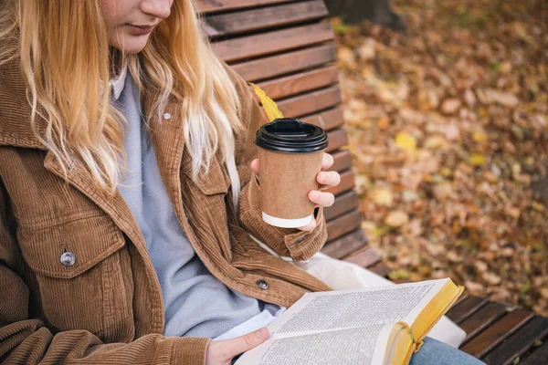 stock image A young woman sits on a bench in an autumn park, drinks coffee and reads a book, close-up.