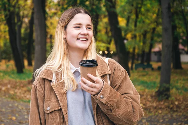 An attractive young woman in a corduroy jacket with a cup of coffee walks in the autumn park.
