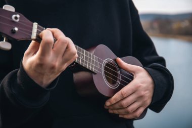 Genç bir adam ukulele gitarını doğada nehrin bulanık arka planında, rahatlama ve müzikal yaratıcılık kavramında çalar..