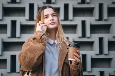 Elinde bir fincan kahveyle telefonda konuşan çekici genç bir kadın geometrik beton arka planda....