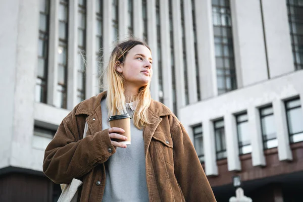 stock image Stylish young woman in a corduroy jacket with a cup of coffee on the background of the university.