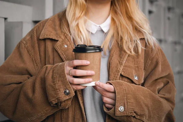 A glass of coffee in a craft disposable glass in the hands of a woman in a corduroy jacket, close-up.