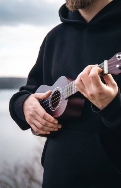 Genç bir adam ukulele gitarını doğada nehrin bulanık arka planında, rahatlama ve müzikal yaratıcılık kavramında çalar..