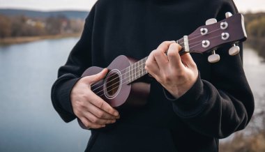 Genç bir adam ukulele gitarını doğada nehrin bulanık arka planında, rahatlama ve müzikal yaratıcılık kavramında çalar..
