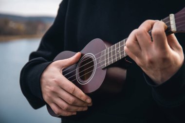 Genç bir adam ukulele gitarını doğada nehrin bulanık arka planında, rahatlama ve müzikal yaratıcılık kavramında çalar..