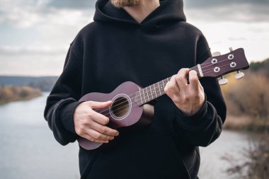 Genç bir adam ukulele gitarını doğada nehrin bulanık arka planında, rahatlama ve müzikal yaratıcılık kavramında çalar..